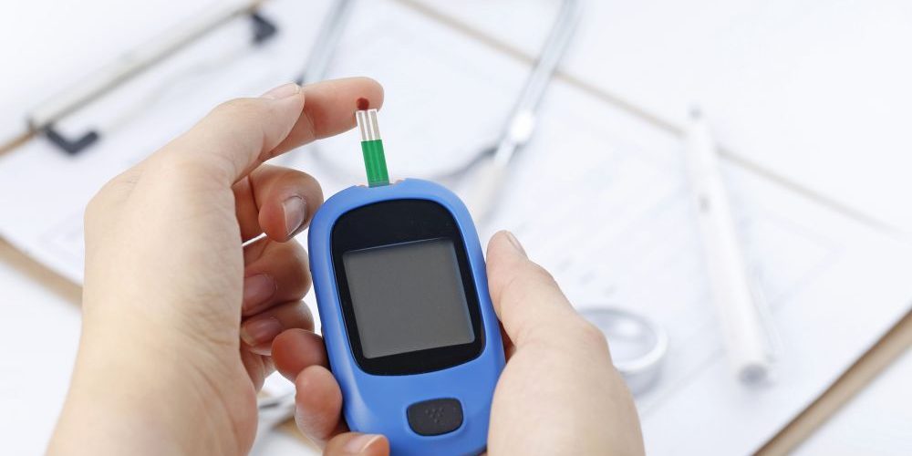 Hand holding a blood glucose meter measuring blood sugar, the background is a stethoscope and chart file